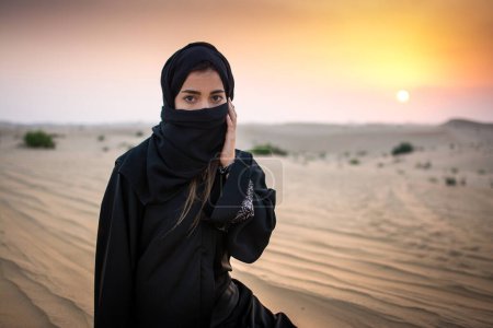 Téléchargez les photos : Portrait d'une jeune femme arabe portant des vêtements noirs traditionnels lors d'un magnifique coucher de soleil sur le désert. - en image libre de droit