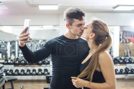 Foto de Hermosa joven pareja deportiva tomando foto selfie en el teléfono inteligente mientras se besa en el gimnasio. - Imagen libre de derechos