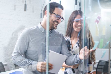 Téléchargez les photos : Femme et homme souriants discutant d'idées et de remue-méninges avec des notes collantes sur le mur de verre au bureau - en image libre de droit