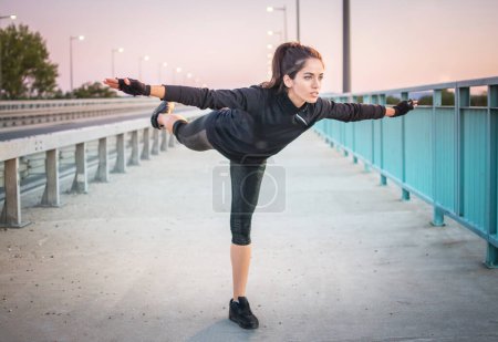 Photo for Healthy young sportswoman doing balance exercise standing on one leg with raised hands outdoors. - Royalty Free Image