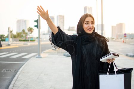 Téléchargez les photos : Belle femme musulmane avec des sacs à provisions, téléphone et portefeuille attelage d'un taxi dans la rue - en image libre de droit