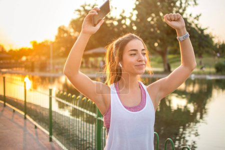 Téléchargez les photos : Joyeuse femme brune portant des vêtements de sport dansant tout en utilisant un smartphone et des écouteurs à l'extérieur. - en image libre de droit