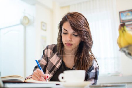 Téléchargez les photos : Portrait d'une étudiante sérieuse faisant ses devoirs tout en prenant un café le matin à la maison - en image libre de droit