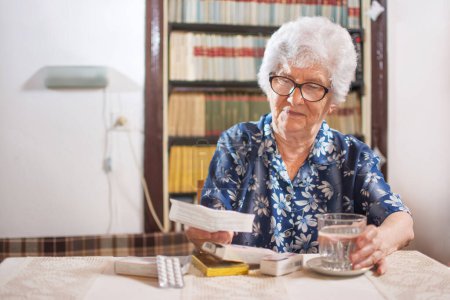 Téléchargez les photos : Femme âgée tenant un verre d'eau et lisant une ordonnance à la maison - en image libre de droit
