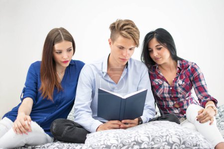 Téléchargez les photos : Trois lycéens lisant ensemble un livre assis sur le lit - en image libre de droit
