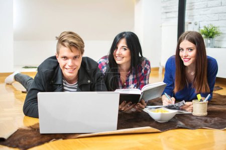 Téléchargez les photos : Groupe de trois jeunes étudiants apprenant avec un ordinateur portable et des livres à la maison - en image libre de droit