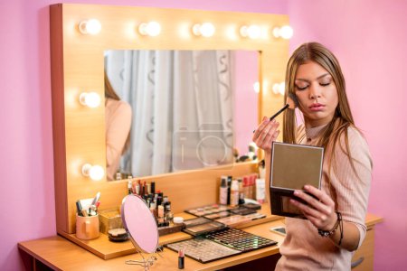 Téléchargez les photos : Jeune femme regardant dans le miroir et appliquant cosmétique avec un pinceau à la maison. - en image libre de droit
