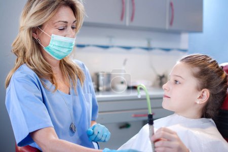 Foto de Chica joven sosteniendo tubo de succión mientras dentista profesional se prepara para trabajar en sus dientes en la clínica dental - Imagen libre de derechos