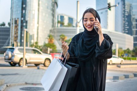 Foto de Mujer joven de Oriente Medio con bolsas de compras hablando por teléfono móvil en la calle de la ciudad - Imagen libre de derechos