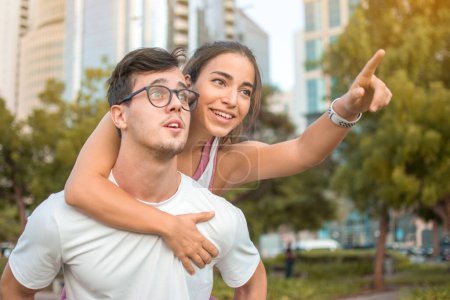 Photo for Happy cheerful caucasian couple in sportswear having piggy back outdoors. Young woman showing something with finger to her boyfriend in the park. - Royalty Free Image