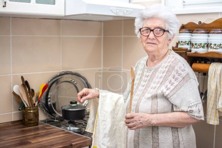 Foto de Mujer mayor con cuchara de mezcla y paño alrededor de su cocina de la mano en la cocina. - Imagen libre de derechos
