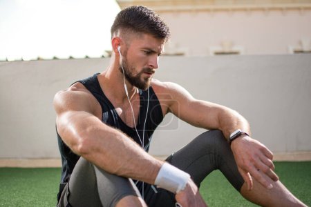 Foto de Hombre barbudo guapo en ropa deportiva con auriculares mirando smartwatch durante el descanso de ejercicio al aire libre. - Imagen libre de derechos