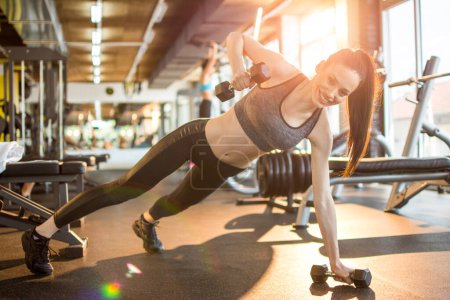 Téléchargez les photos : Femme de remise en forme en vêtements de sport faisant des pompes à une main avec des poids à la salle de gym. - en image libre de droit