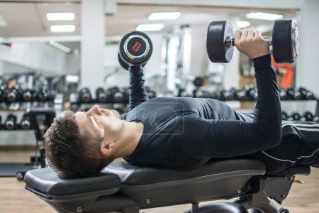 Photo for Handsome young man lying on exercise bench and lifting dumbbells at gym. - Royalty Free Image
