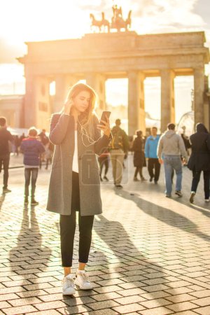 Téléchargez les photos : Portrait intégral d'une jeune femme avec écouteurs écoutant de la musique au téléphone devant la porte de Brandebourg à Berlin, Allemagne. - en image libre de droit