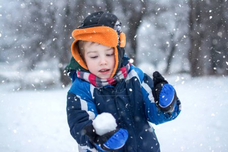 Téléchargez les photos : Portrait d'adorable petit garçon jouant avec boule de neige pendant la journée d'hiver enneigée - en image libre de droit