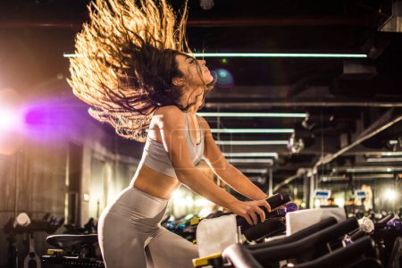 Foto de Vista lateral de la atractiva mujer joven con el pelo largo en el aire durante el entrenamiento de ciclismo en el gimnasio. - Imagen libre de derechos