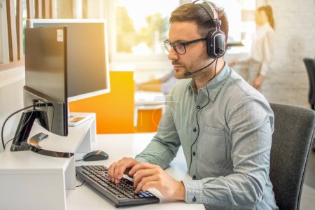 Photo for Male professional call center telesales agent wearing headset using computer in customer care support service office. - Royalty Free Image