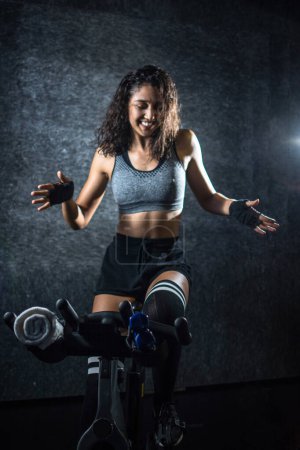 Cheerful sporty woman dancing on exercise bike during cycling ride indoors.