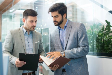 Photo for Businessmen working together with documents in office. Handsome male partners discussing paperwork while standing in office lobby. - Royalty Free Image