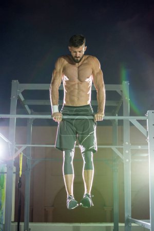 Photo for Athletic handsome man doing dip stand exercise on horizontal bar during calisthenics outdoors at night. Strength and endurance exercise. - Royalty Free Image