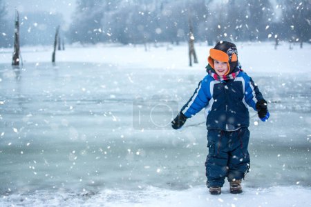 Téléchargez les photos : Joyeux garçon debout près de la rivière gelée le jour neigeux - en image libre de droit