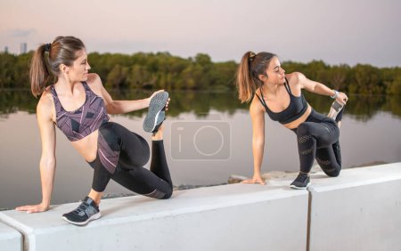 Zwei junge, sportliche Frauen, die sich gemeinsam im Freien strecken. Fitness-Frauen in einbeinigen Königstauben-Posen am Fluss