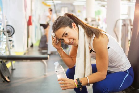 Chica fitness cansada con toalla blanca en el cuello y botella de agua en un gimnasio.