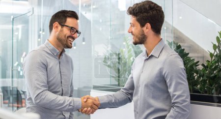 Foto de Dos hombres de negocios sonrientes estrechando las manos mientras están de pie en la oficina moderna. - Imagen libre de derechos