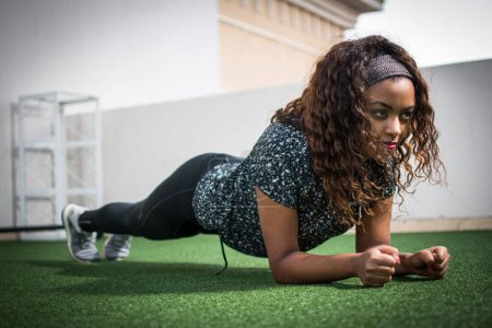 Téléchargez les photos : Belle femme afro-américaine en vêtements de sport démontrant l'exercice de planche pour la force abdominale. - en image libre de droit