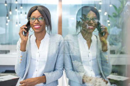 Foto de Retrato de una mujer sonriente hablando por teléfono celular mientras se apoya en la pared de cristal en la oficina. - Imagen libre de derechos