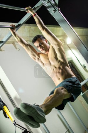 Photo for Handsome shirtless man doing pull ups in the L-sit position on horizontal bar outdoors at night. Calisthenics training. - Royalty Free Image