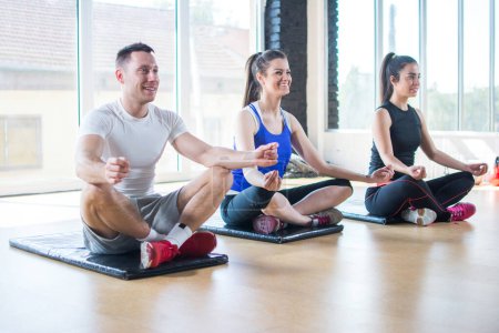 Téléchargez les photos : Groupe de jeunes amis sportifs pratiquant le yoga, assis jambes croisées sur le sol en position lotus à la salle de gym - en image libre de droit