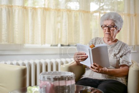 Foto de Mujer mayor leyendo libro y relajándose en sillón en casa. - Imagen libre de derechos