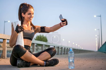 Téléchargez les photos : Souriant fille sportive prenant un autoportrait au téléphone après l'entraînement de nuit - en image libre de droit