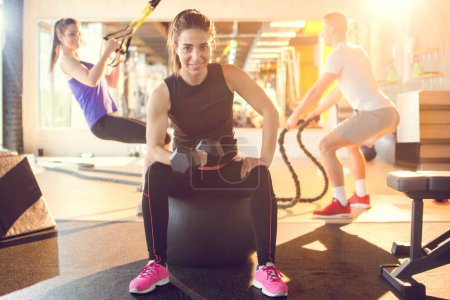 Photo for Young sporty woman lifting weights at gym with her friends in the background - Royalty Free Image