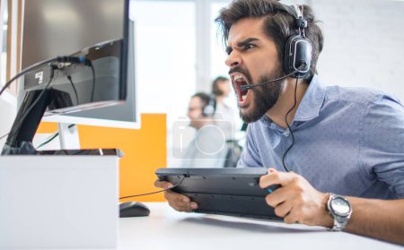 Excited handsome male customer support operator or game tester with headset shouting on computer monitor and holding keyboard at office