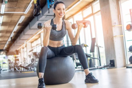 Foto de Mujer de fitness en entrenamiento de ropa deportiva con pesas mientras está sentada en la pelota suiza en el gimnasio
. - Imagen libre de derechos