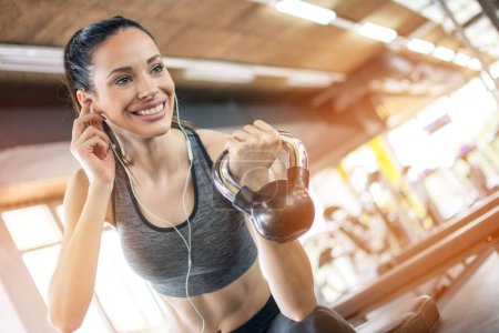 Photo for Low angle view of smiling fit woman with earphones lifting kettlebell during morning workout in fitness gym. - Royalty Free Image