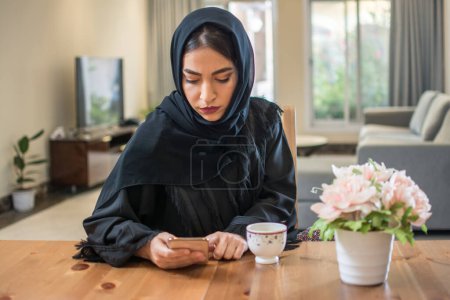 Foto de Mujer musulmana joven usando hijab usando el teléfono en casa. - Imagen libre de derechos