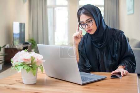 Téléchargez les photos : Jolie jeune femme du Moyen-Orient portant hijab et lunettes à l'aide d'un ordinateur portable à la maison. - en image libre de droit