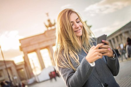 Téléchargez les photos : Jeune femme souriante utilisant le téléphone devant la porte de Brandebourg, Berlin, Allemagne. - en image libre de droit