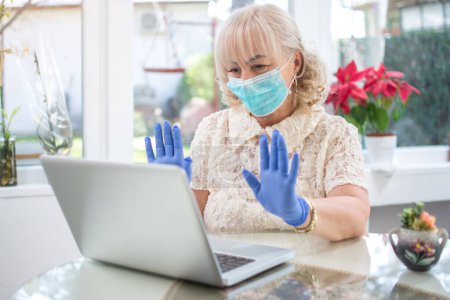 Photo for Senior woman wearing protective mask, showing hands in latex gloves during video call with family. Staying at home, Coronavirus, quarantine, senior and technology concept. - Royalty Free Image