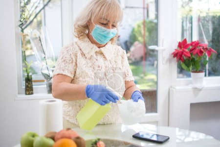 Photo for Mid age woman wearing protective mask and gloves disinfecting her phone with spray disinfectant liquid and paper tissue against the virus at home - Royalty Free Image