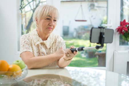 Foto de Mujer mayor que usa selfie stick monopod para hacer videollamadas o fotos de selfie en su teléfono inteligente en casa. - Imagen libre de derechos