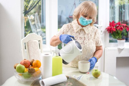 Téléchargez les photos : Femme d'âge moyen en gants de protection et masque médical préparant le thé dans la cuisine. Prévention du virus Corona. Reste à la maison.. - en image libre de droit