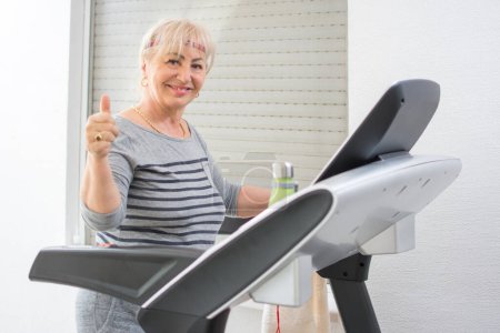 Photo for Active middle aged woman in sportswear showing thumb up on treadmill at home. - Royalty Free Image
