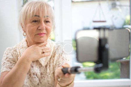 Foto de Mujer madura sosteniendo palo selfie con teléfono inteligente mientras tiene una videollamada en casa - Imagen libre de derechos