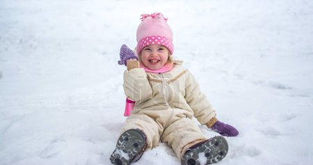Foto de Niña alegre sentada en la nieve durante el frío día de invierno - Imagen libre de derechos