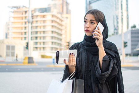Téléchargez les photos : Charmante femme musulmane en abaya parlant sur un téléphone portable dans la rue de la ville - en image libre de droit
